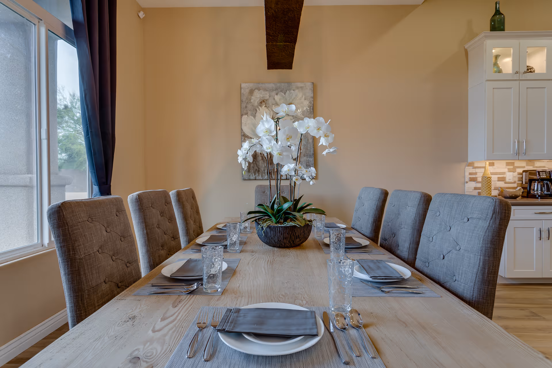 A long dining table with upholstered chairs, a central orchid arrangement, and a kitchen backdrop.