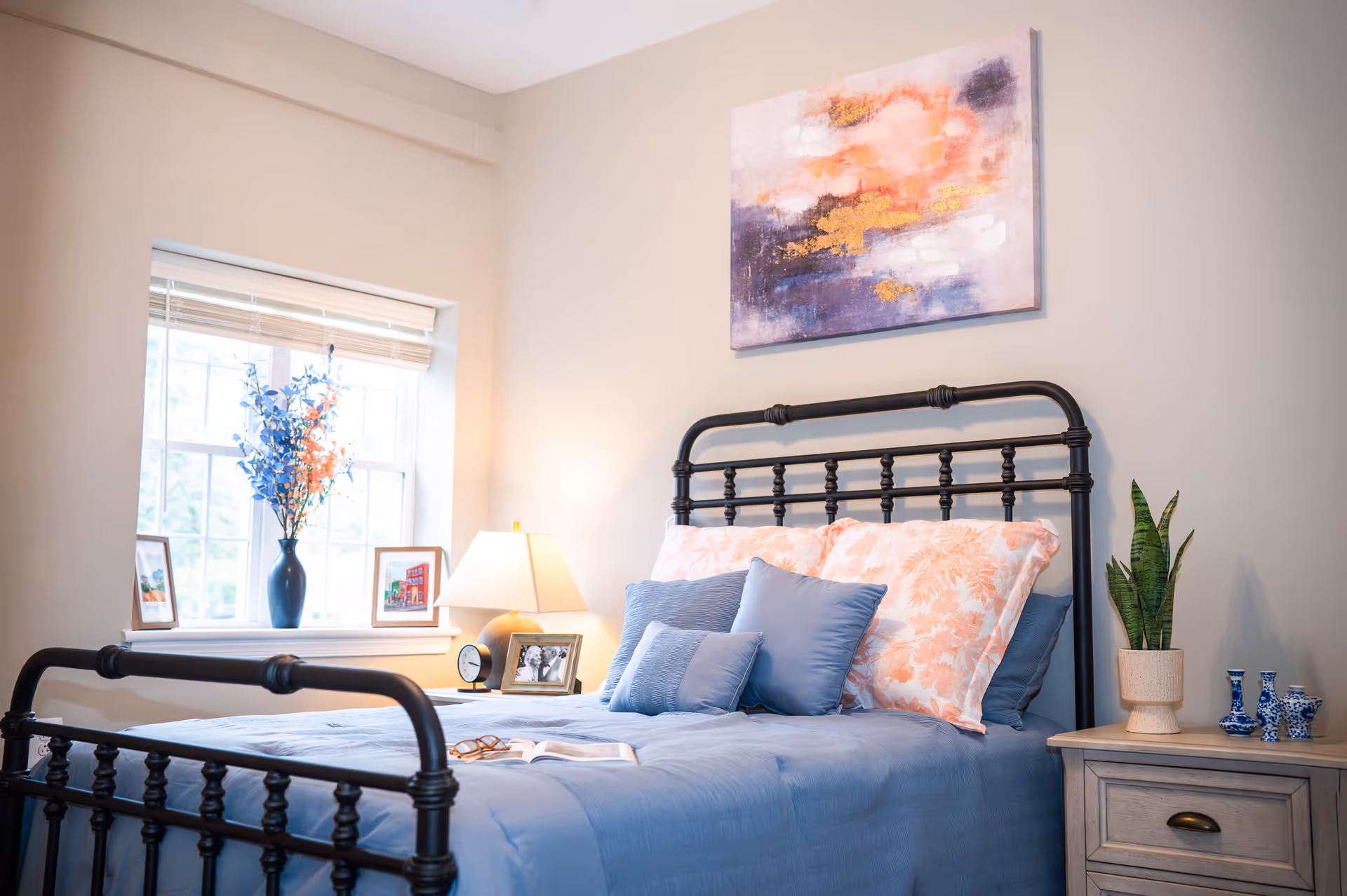 Cozy bedroom with blue bedding, metal bed frame, nightstand, lamp, plant, and window light.
