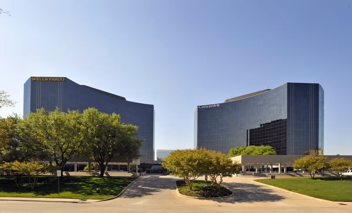 Two curved glass office towers with Wells Fargo and Concentra signage, trees, and a sunny driveway.