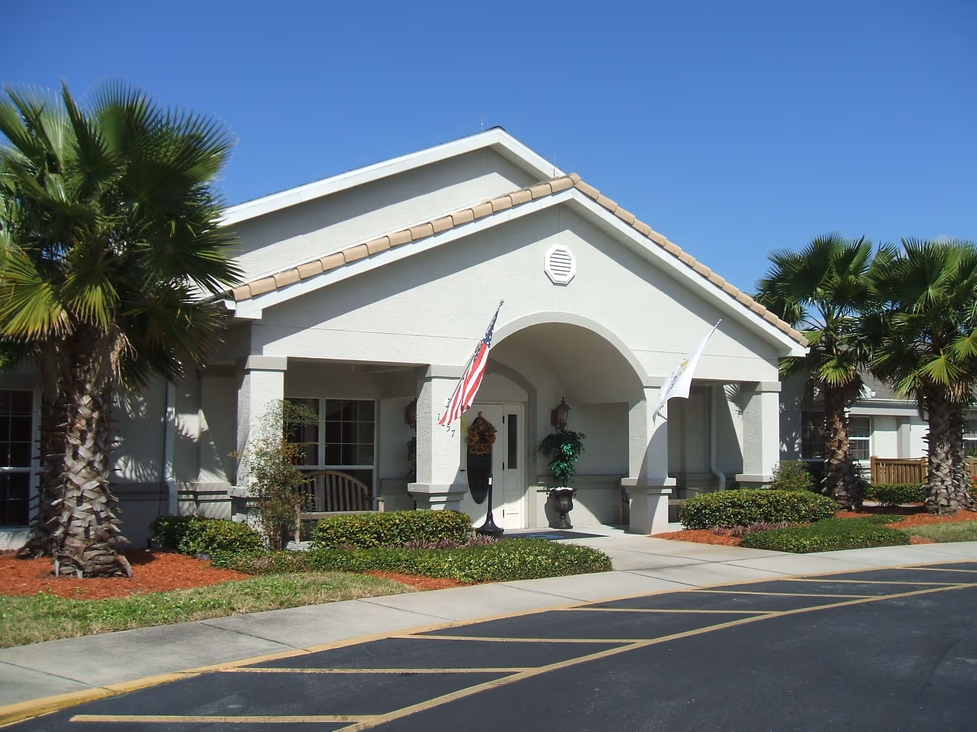 Front exterior view of a single-story building with a covered entrance supported by columns. Two flags, including an American flag, are displayed near the entrance. The building is surrounded by palm trees, shrubs, and well-maintained landscaping under a clear blue sky.