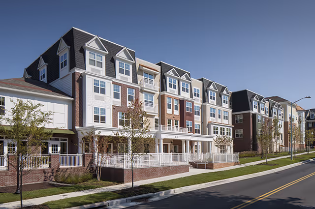 Front exterior of a multi-story senior living building with brick and white siding, porches, and dormer windows along a street.