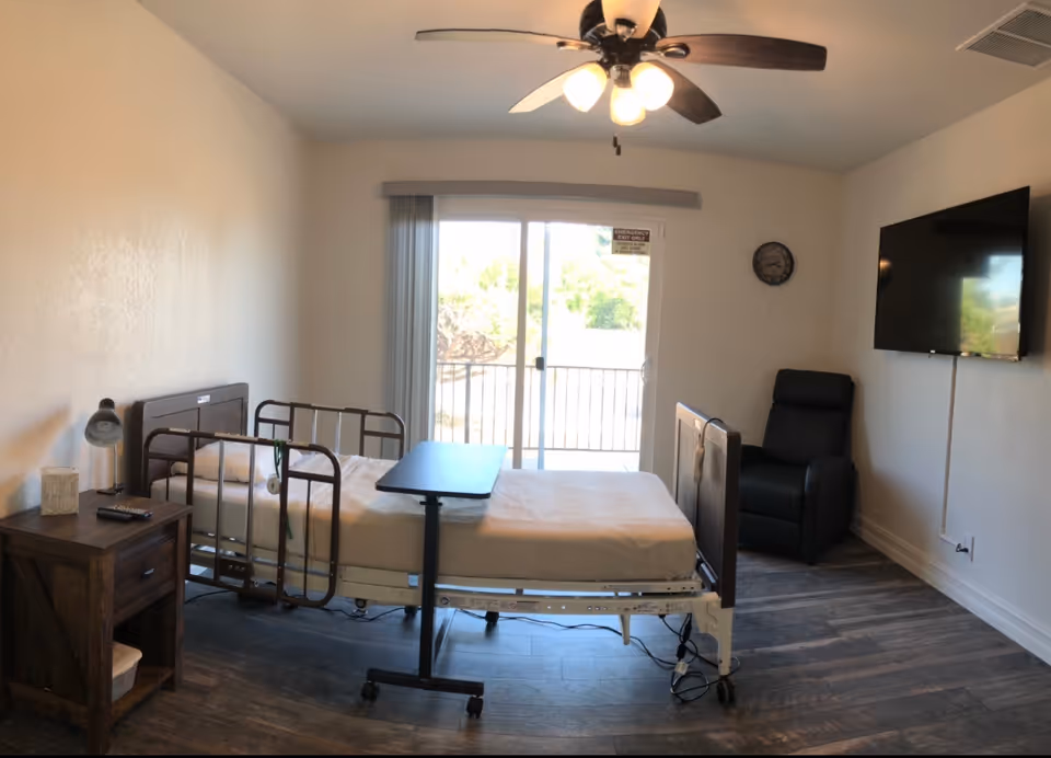 A bedroom in a senior living facility featuring a hospital bed, TV, recliner, and sliding door.