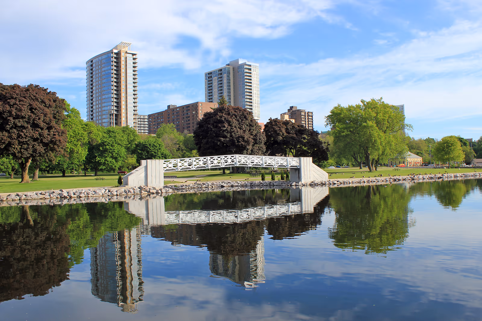Lakeside park with a white footbridge, trees, and reflected city skyline.