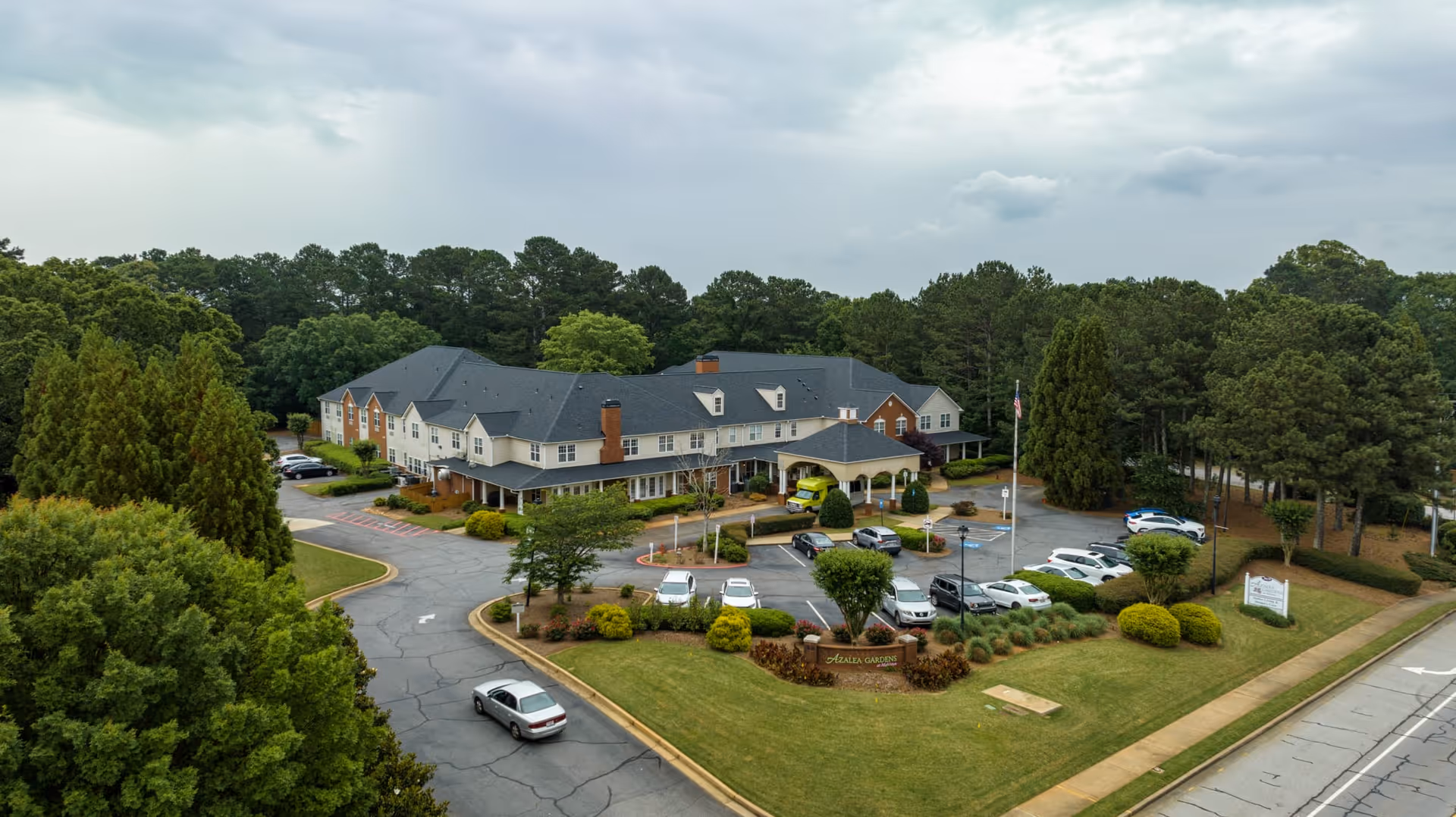 Aerial view of Azalea Gardens Mableton, a large senior living facility surrounded by trees and greenery. The building has a dark roof and light-colored walls with multiple windows. There is a parking lot with several cars parked, a driveway leading to the entrance, and landscaped bushes and grass around the property.