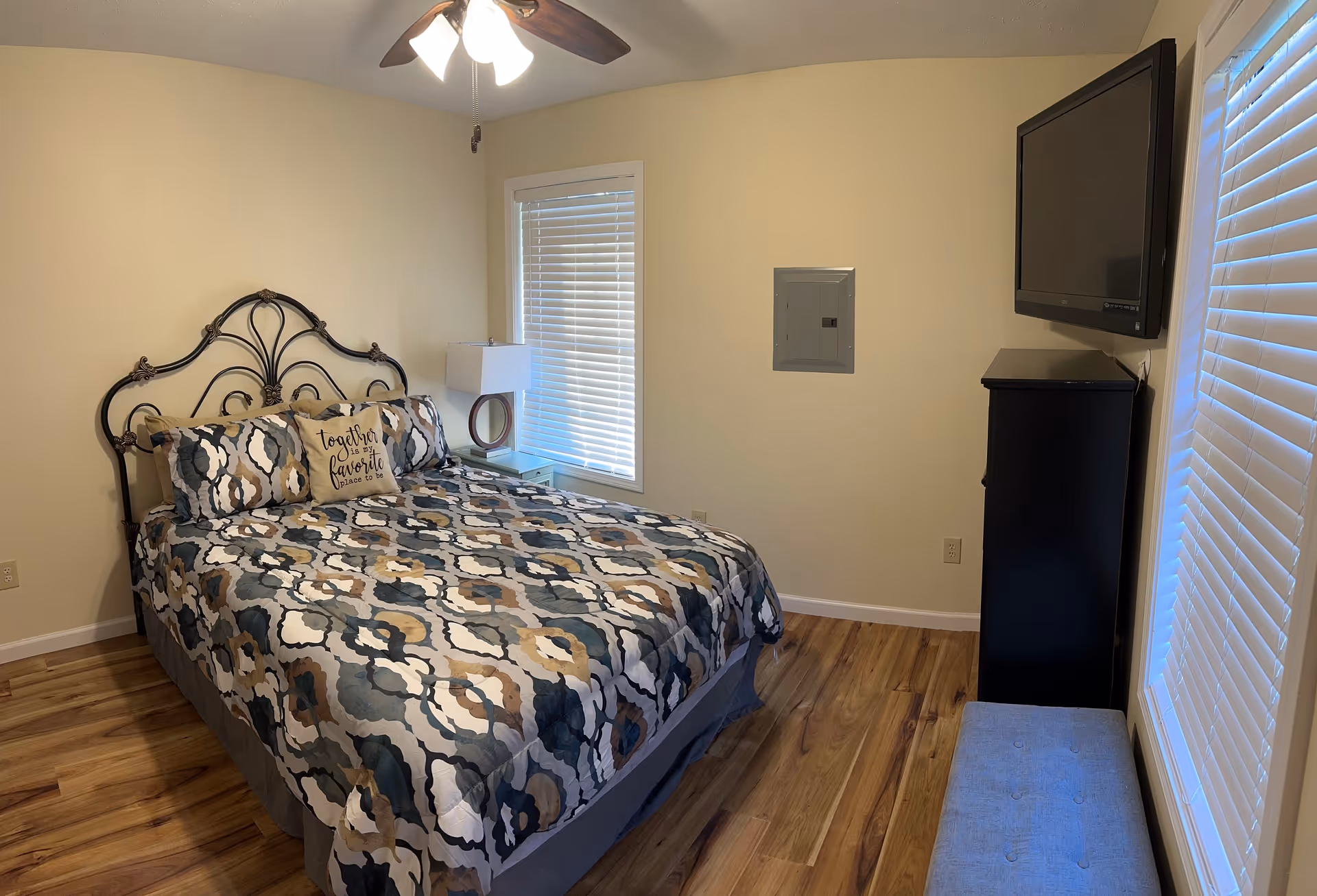 Bedroom with ornate metal headboard bed, patterned quilt, ceiling fan, blinds, and wall-mounted TV.