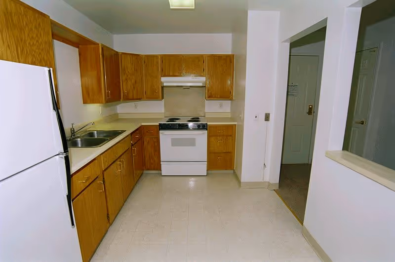 A clean kitchen with wooden cabinets, a white refrigerator on the left, a double sink, a white stove with an exhaust hood, and light-colored countertops and flooring. There is an open doorway leading to another room on the right.