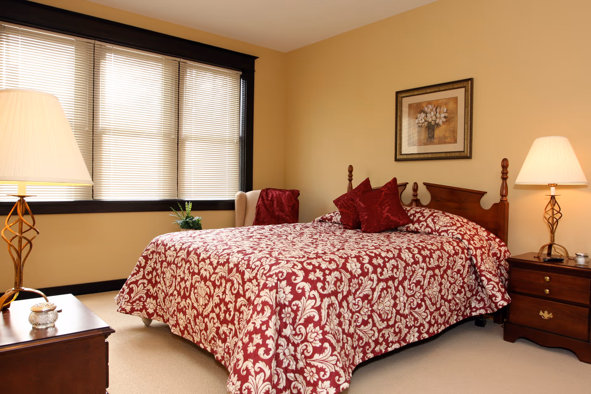 A cozy bedroom with a large bed covered in a red and white patterned bedspread and matching pillows. The room has beige walls, a window with blinds, two wooden nightstands with lamps, a small plant, and a framed floral picture on the wall above the bed.