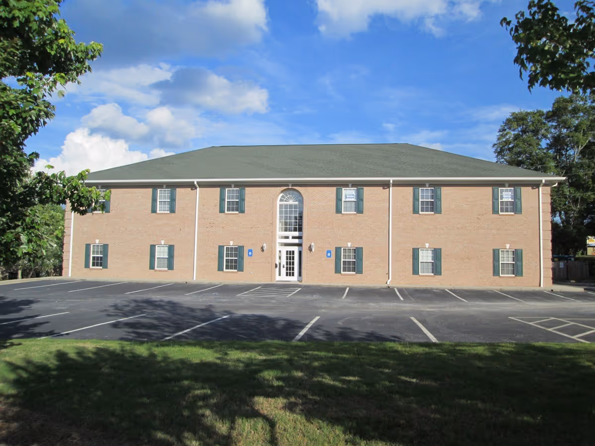 Brick two-story building with green shutters, a central arched entry, and a spacious parking lot.