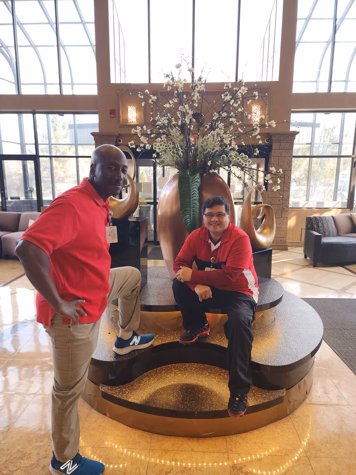 Two men wearing red shirts posing inside a spacious, well-lit lobby area with large windows. One man is standing with one foot on a step, and the other is sitting on a circular platform with a large decorative vase and floral arrangement behind them. The lobby has comfortable seating and a polished floor.