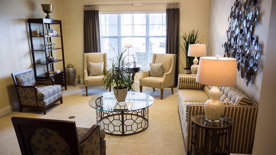 A stylish senior living lounge with beige seating, coffee table, plants, and wall decor.