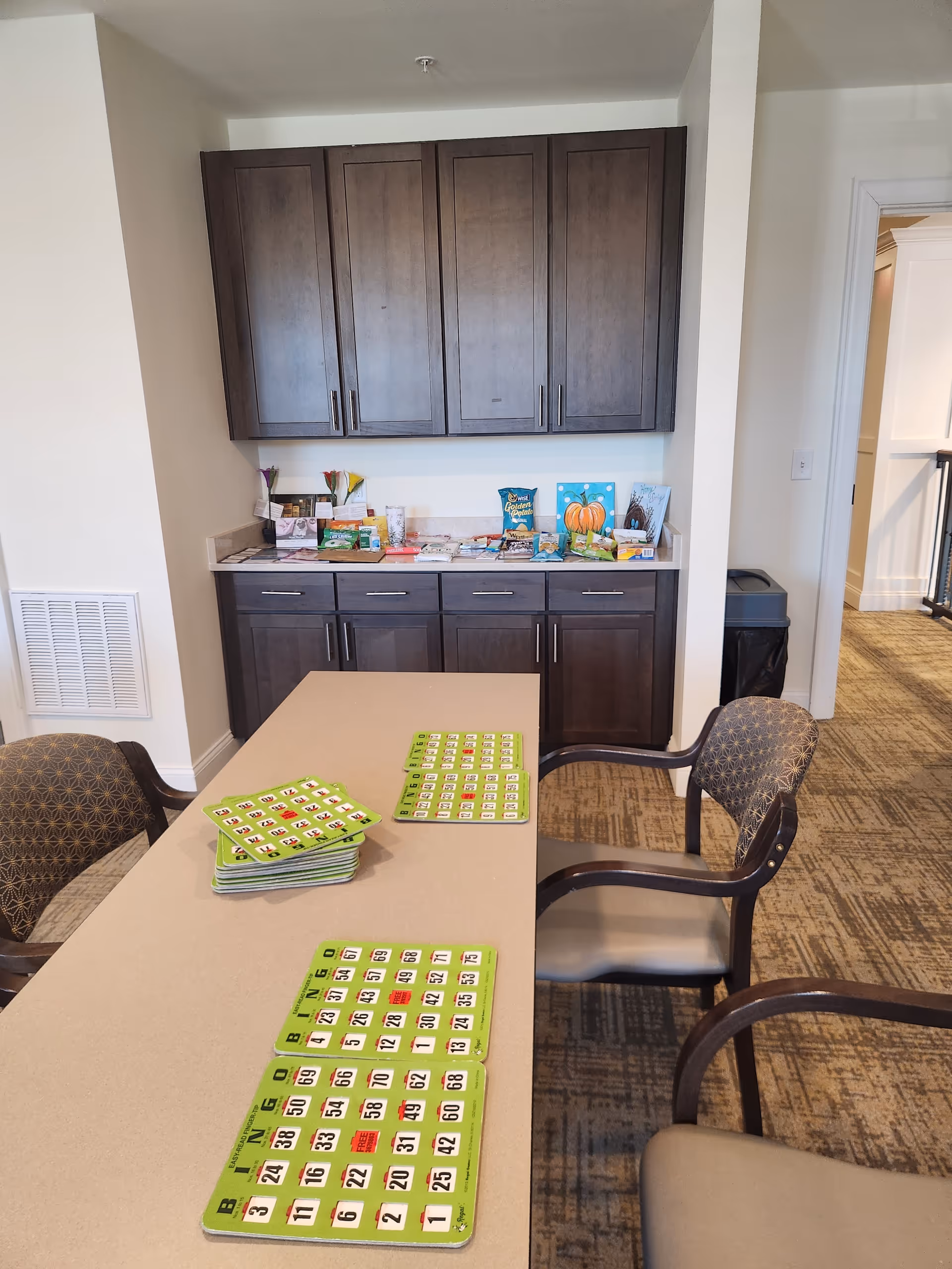Table with bingo boards and snacks in a senior living facility common room.