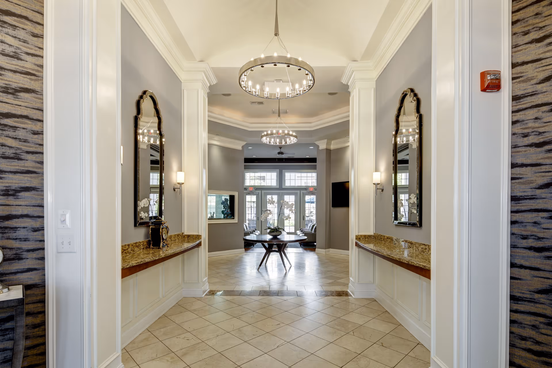 A bright and elegant hallway with tiled floors and light gray walls featuring two large decorative mirrors above granite countertops on either side. The hallway leads to a common area with large windows and glass doors letting in natural light. Two modern circular chandeliers hang from the ceiling, and there are wall sconces providing additional lighting.