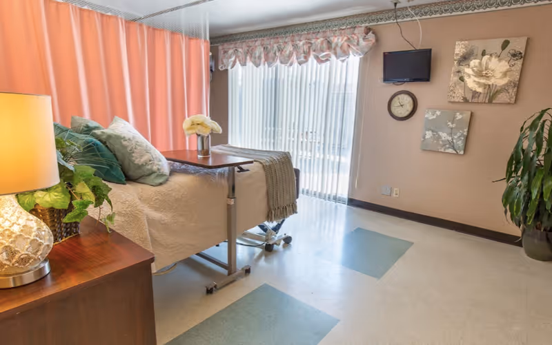 A care facility bedroom with a hospital-style bed, overbed table, lamp, potted plants, wall art and a wall-mounted TV by sliding blinds.