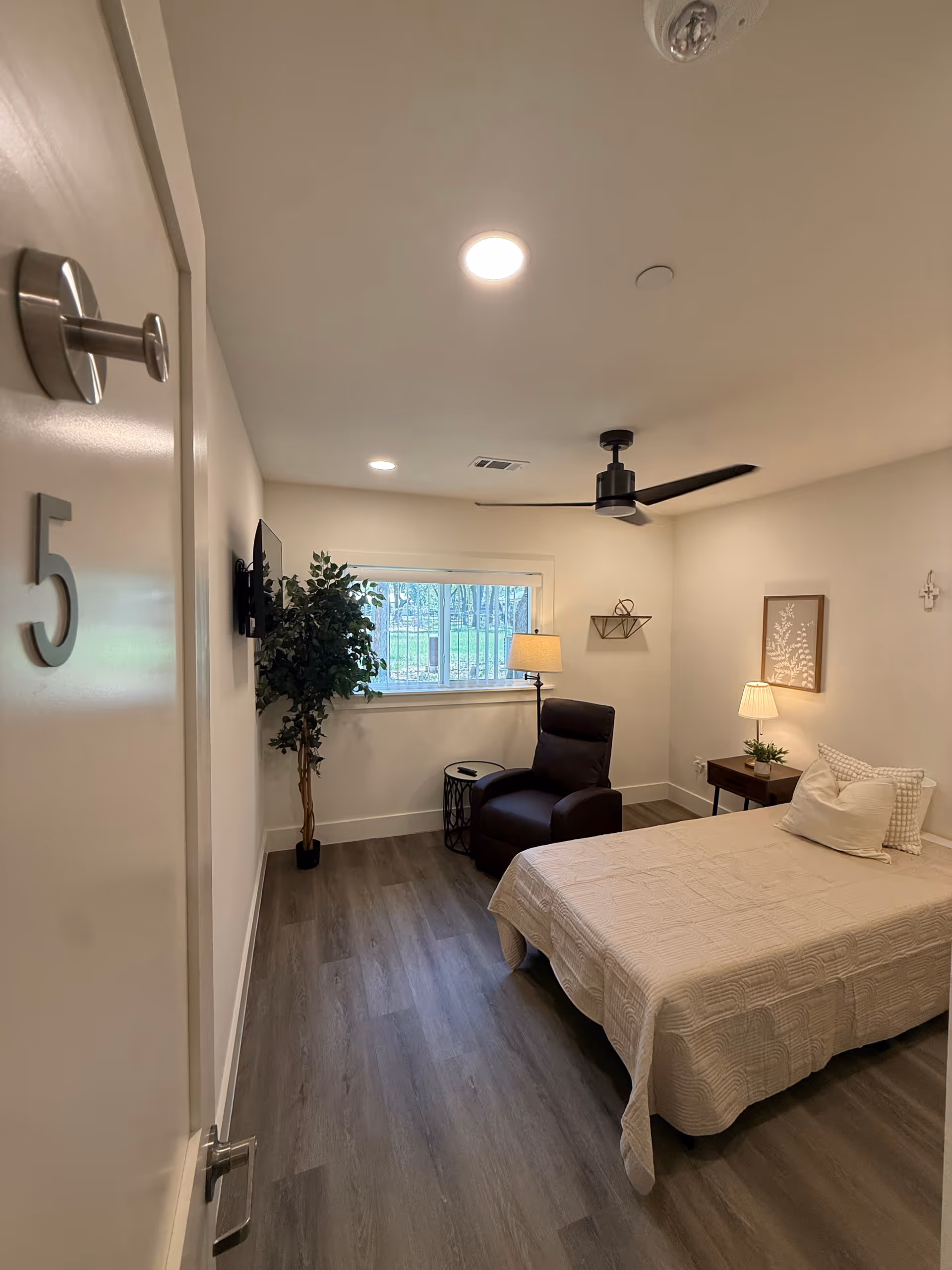 A cozy bedroom with a bed, recliner, nightstand, plant, and window in a senior living unit.