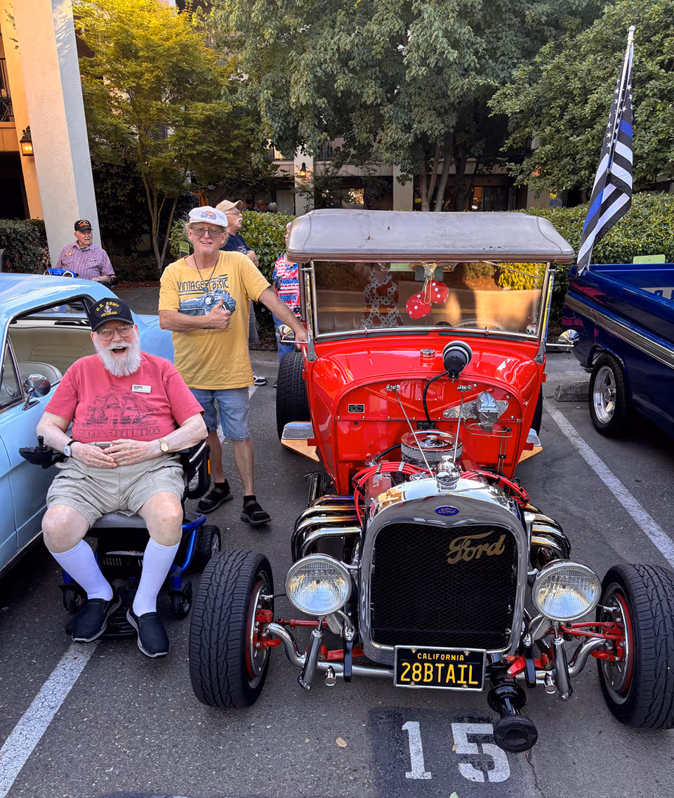 Two elderly residents stand beside a bright red vintage Ford in a parking lot.