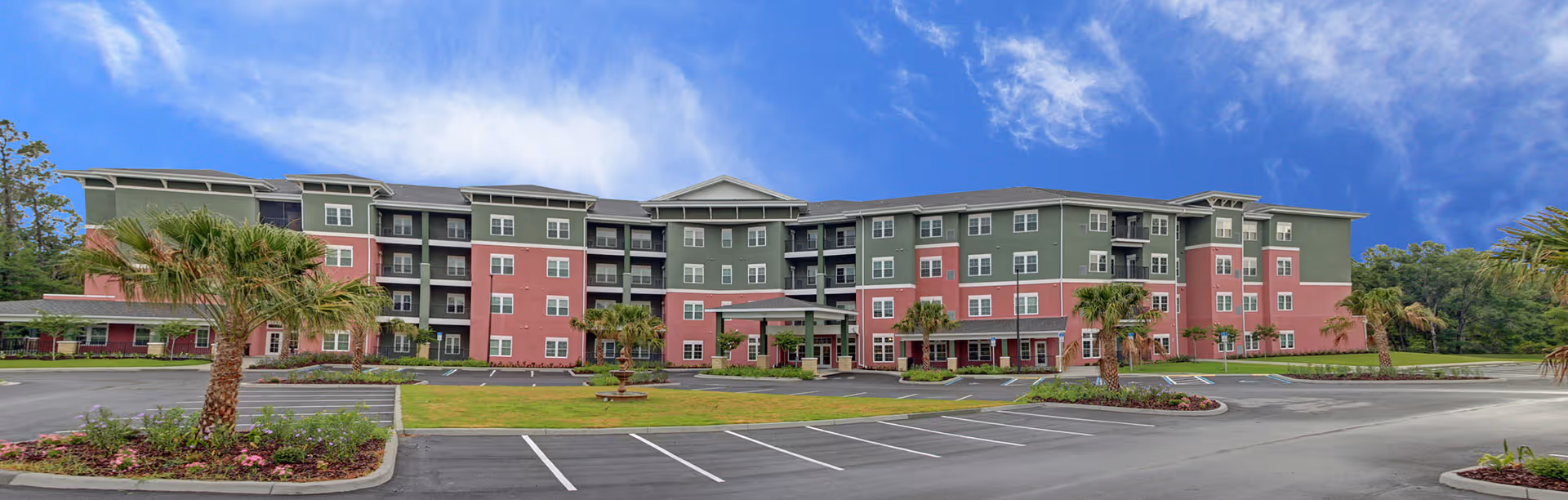 Wide exterior view of a multi-story senior living facility building with green and pink walls, surrounded by palm trees and landscaping, under a partly cloudy blue sky. There is a parking lot in front with marked parking spaces and a circular driveway with a small fountain in the center.