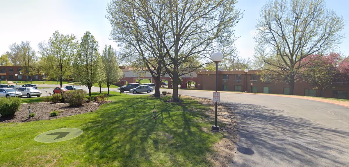 Exterior view of a senior living campus with trees, lawn, and parked cars.