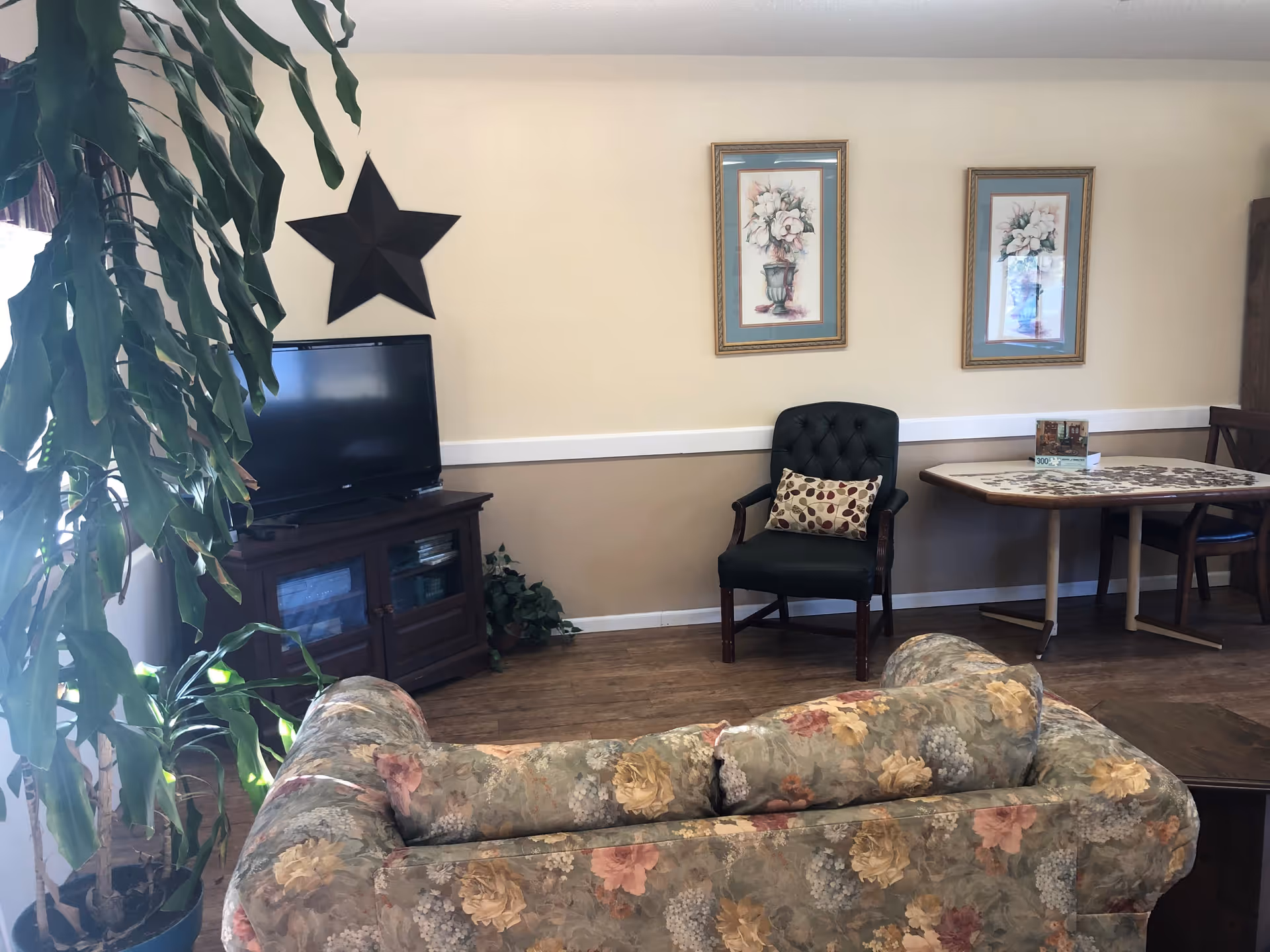 Communal living room with a floral sofa in the foreground, a TV on a wooden cabinet, an armchair and a small table beneath framed artwork on a beige wall.