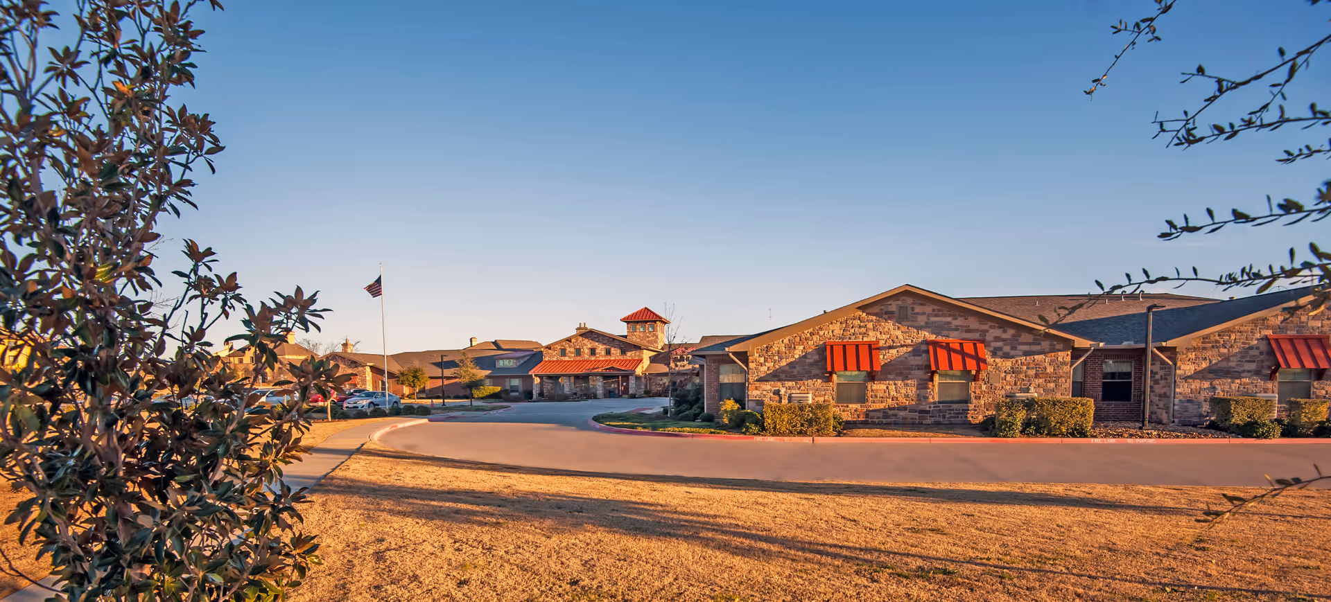 Sunlit exterior of Willow Bend Assisted Living & Memory Care with stone facade and red awnings.