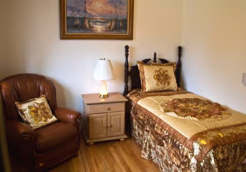 Small bedroom with a single bed dressed in gold-brown bedding, a bedside table with a lamp, an upholstered armchair, and a framed painting on the wall.