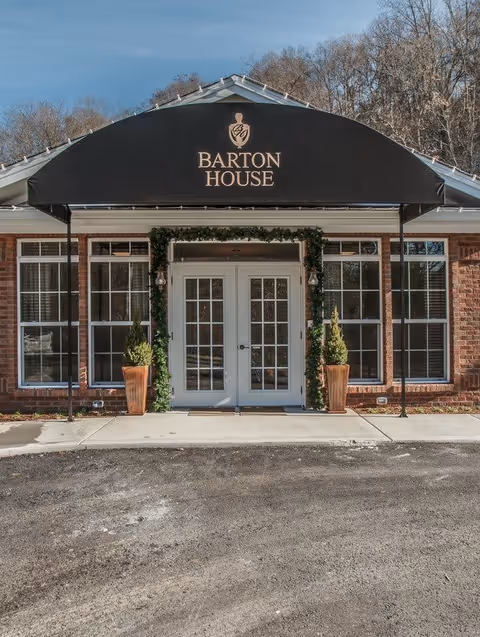 Front entrance of Barton House Memory Care facility with a black awning displaying the facility's name and logo above double glass doors framed by greenery and two potted plants on either side, set against a brick building with large windows.