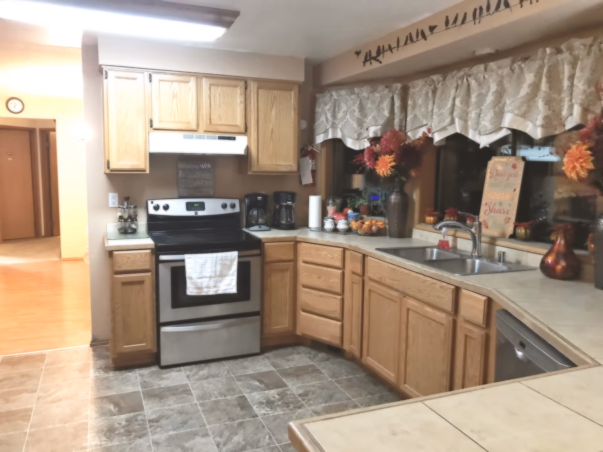 L-shaped kitchen with wooden cabinets, stainless stove, sink, and autumn decor near the window.