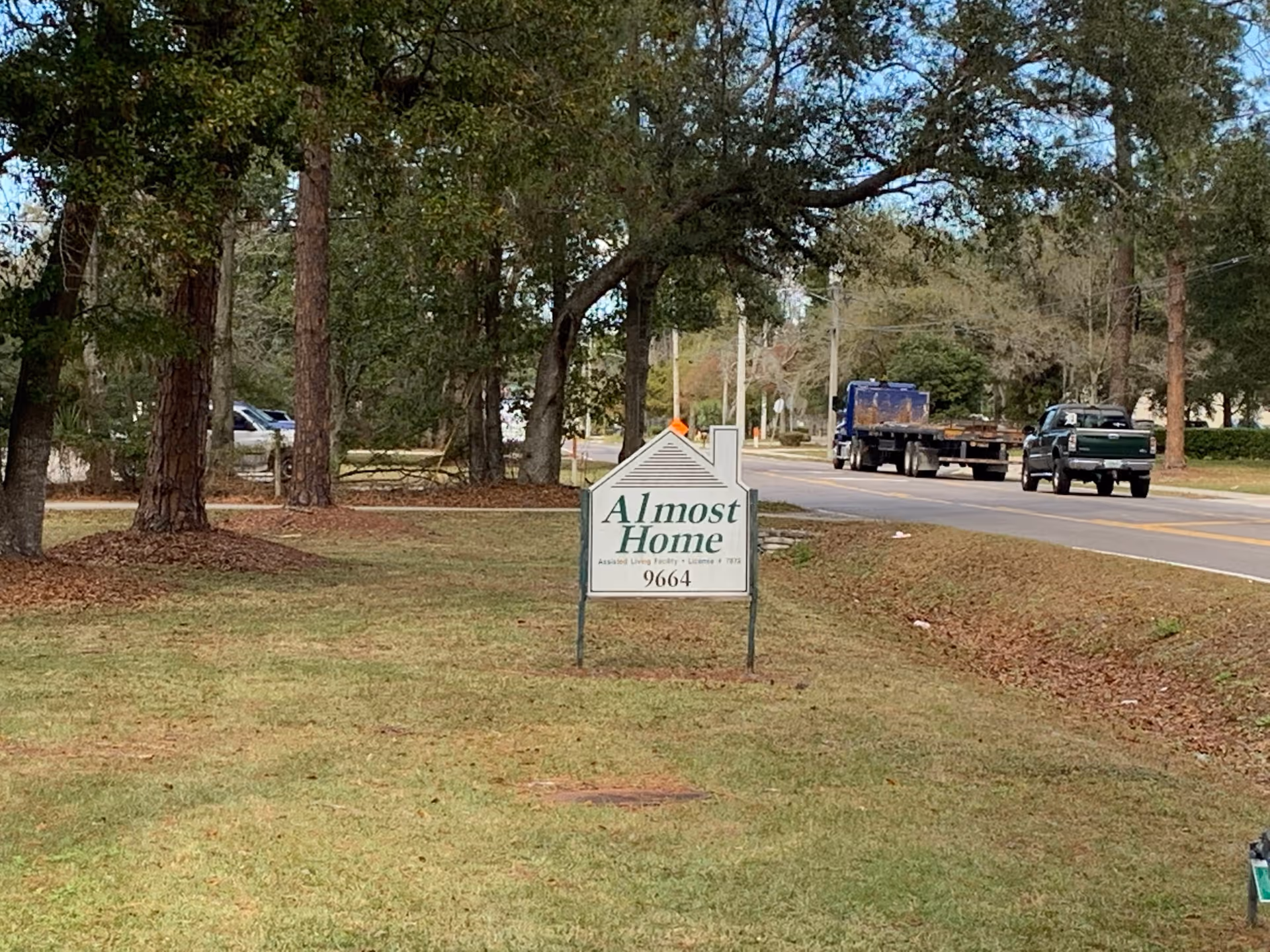A roadside sign for Almost Home Senior Services Inc, an assisted living facility, standing on a grassy area with trees in the background and a road with vehicles passing by.