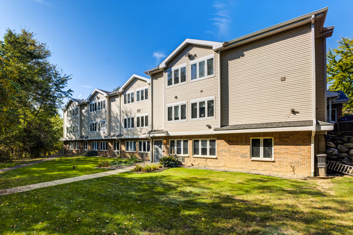 A Dimensions Living Stoughton senior living building with beige siding and brick base, surrounded by lawn.