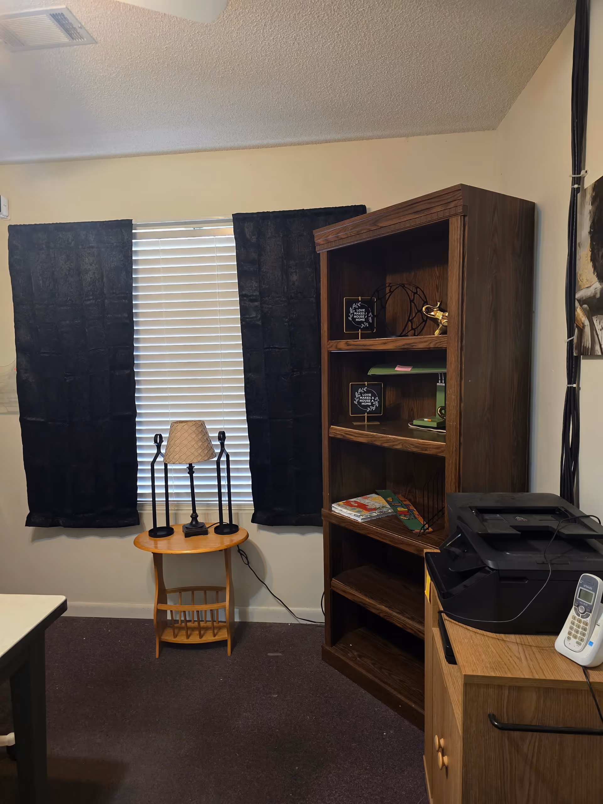 Interior room featuring a tall bookshelf, printer, and a round table with lamps by a window.