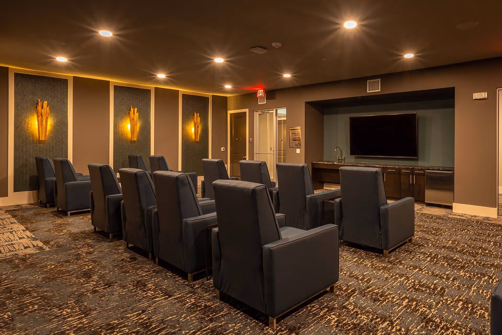 A small theater-style media room with rows of dark reclining chairs facing a large wall-mounted TV and warm wall sconces.