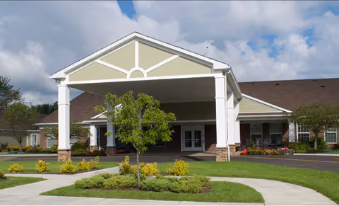 Front exterior view of Orchard Heights facility with a covered entrance, a small tree, landscaped bushes, and a clear sky with some clouds.