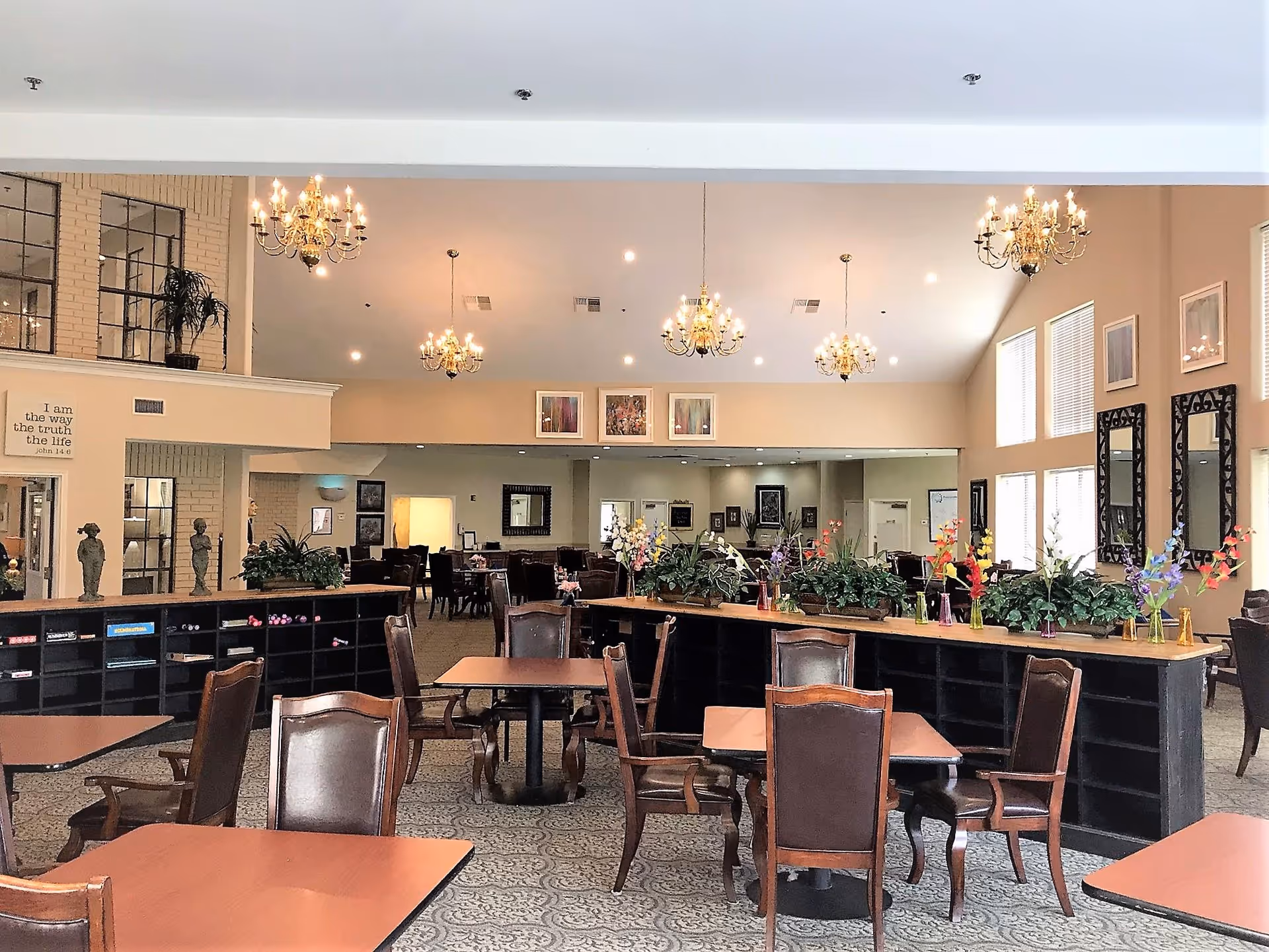 A spacious and well-lit dining area in a senior living facility with multiple wooden tables and chairs arranged neatly. The room features chandeliers hanging from the ceiling, large windows allowing natural light, decorative plants and flowers on a central divider, and framed artwork on the walls.