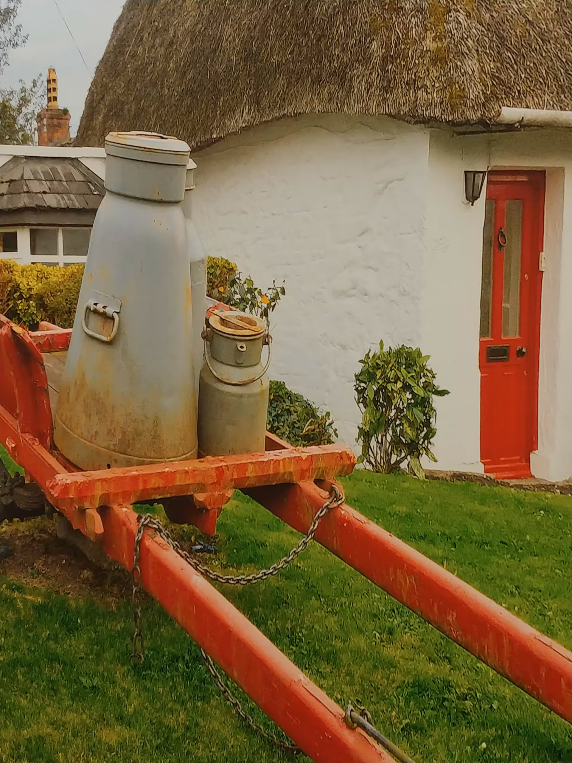 Rustic white building with a thatched roof, red door, and a rusty wagon with milk cans.