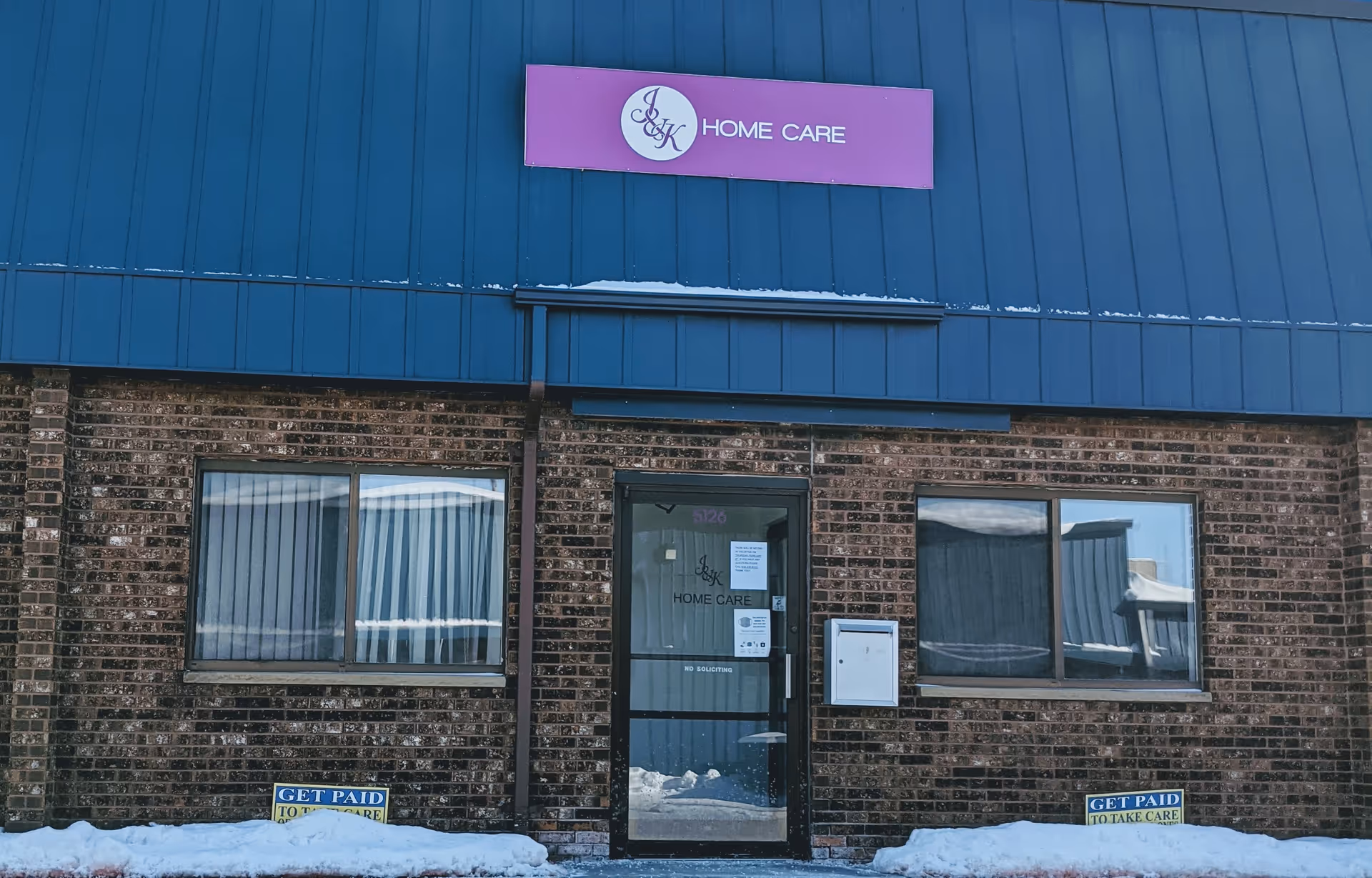 Blue metal facade above brick entrance with purple J&K Home Care sign and snow nearby.