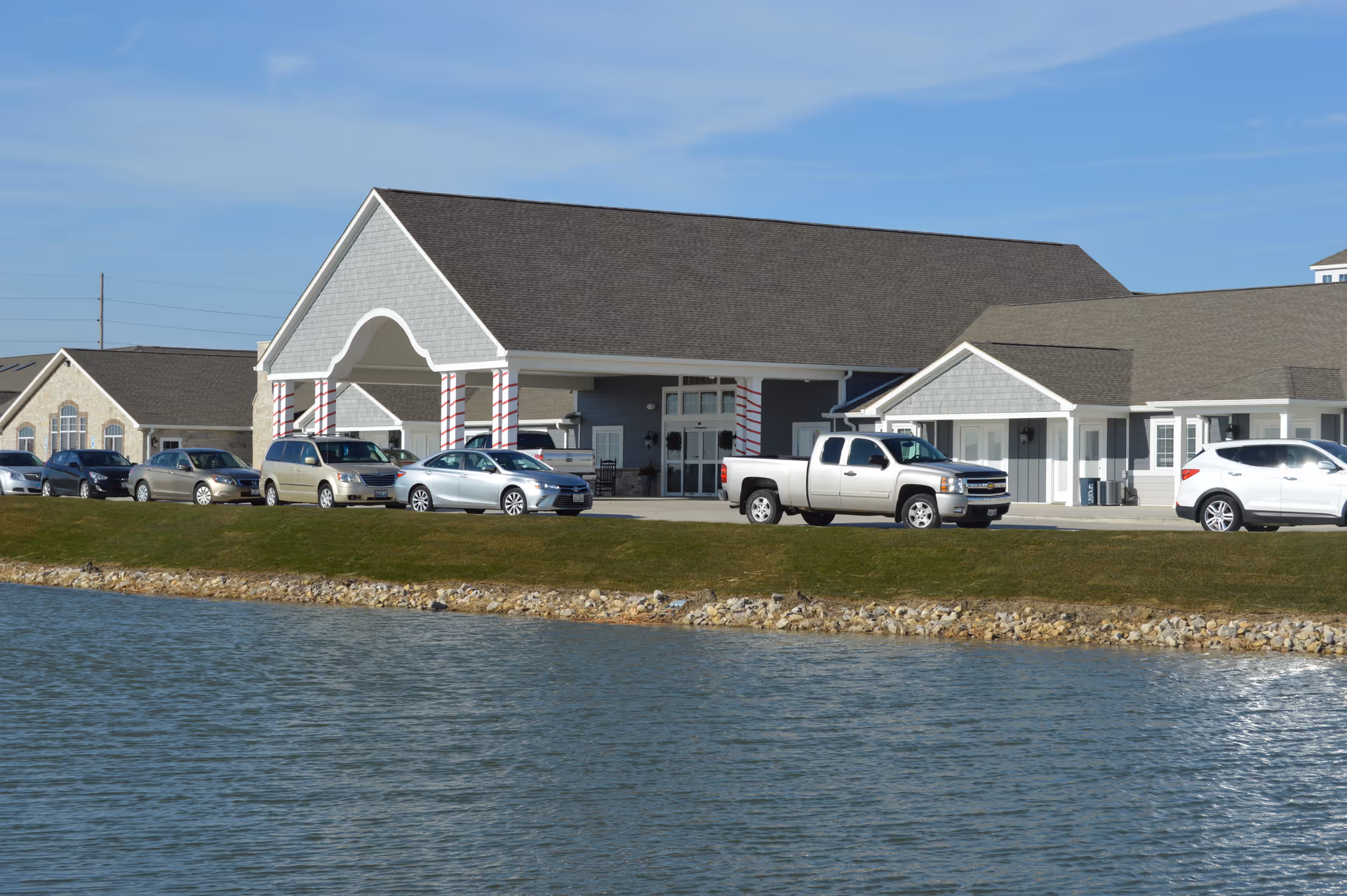 Exterior view of Villas of Holly Brook Assisted Living & Memory Care facility in Rantoul, IL, showing a large building with a covered entrance supported by columns wrapped in red and white stripes. Several cars are parked in front of the building, and a body of water with a rocky shoreline is visible in the foreground under a clear blue sky.