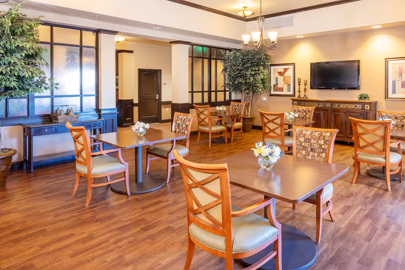 A well-lit dining room with wooden floors, several wooden tables and chairs with cushioned seats, decorative plants, framed artwork on the walls, and a flat-screen TV mounted above a wooden sideboard.