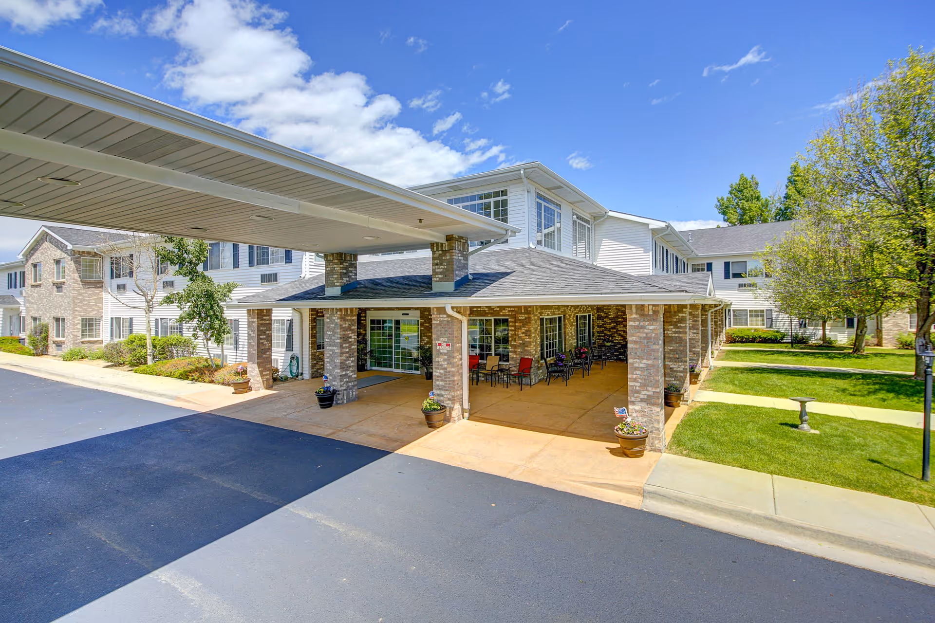 Entrance of Waneka Park Assisted Living facility showing a covered drop-off area with brick pillars, outdoor seating with tables and chairs, a paved driveway, and a well-maintained lawn with trees and a birdbath under a partly cloudy blue sky.