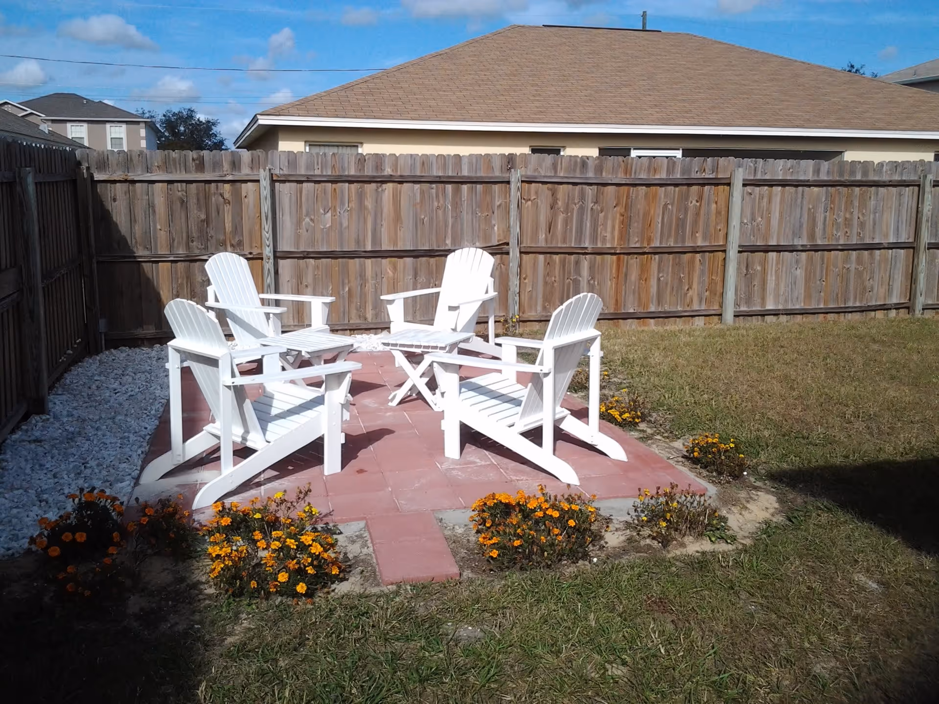 Small fenced backyard patio with four white Adirondack chairs around a small table on red pavers and surrounding flowerbeds.