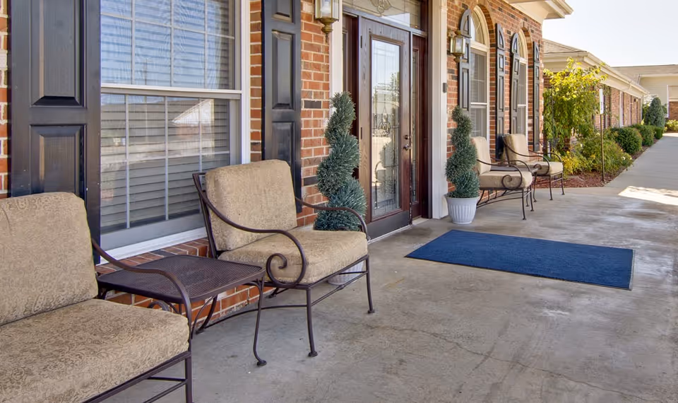 Front entrance of a brick senior living building with cushioned metal chairs, potted topiaries, and a blue welcome mat on a concrete walkway.