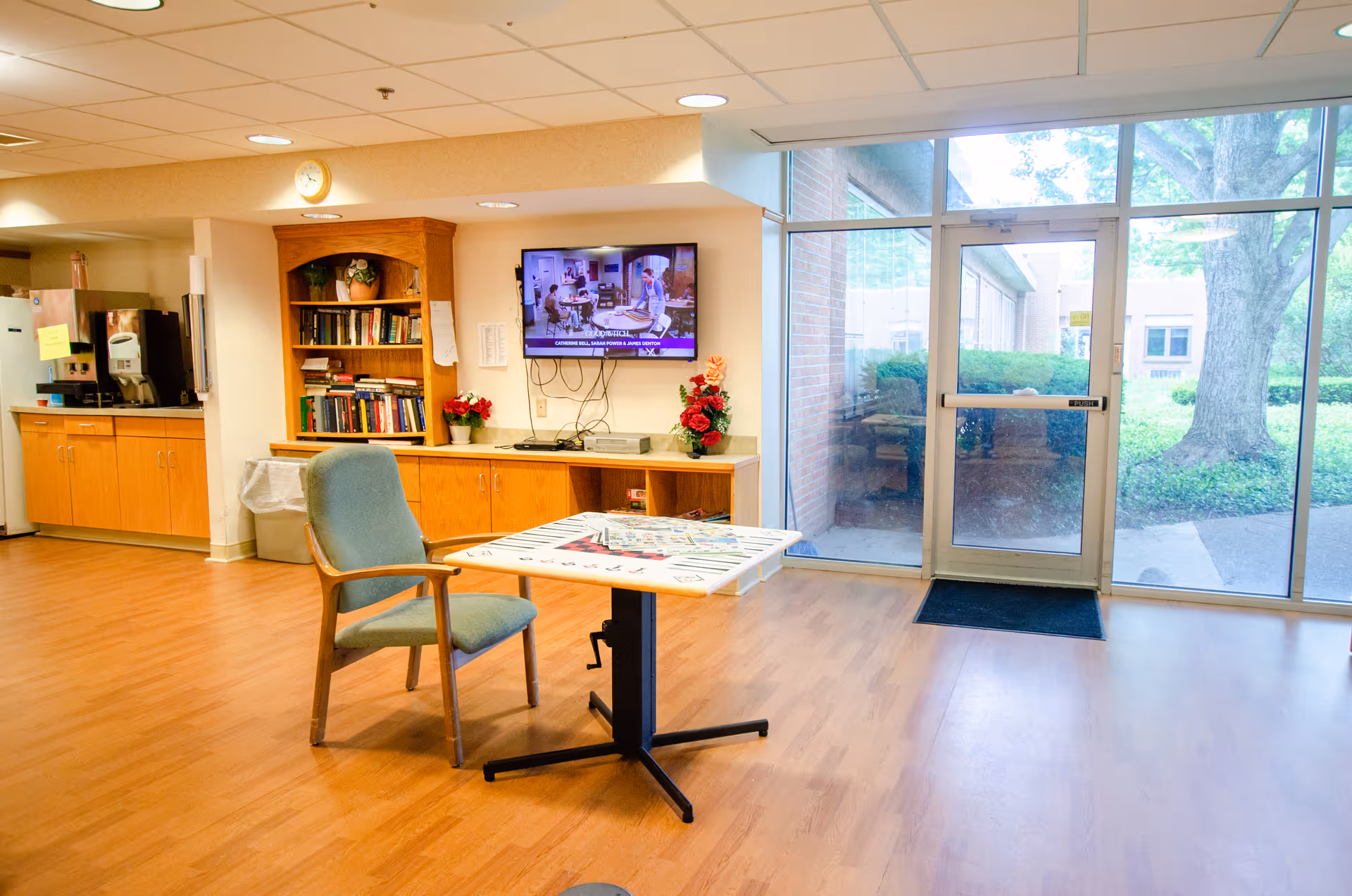 A bright common area in a senior living facility with a single chair and a table featuring a board game. There is a wooden bookshelf filled with books, a wall-mounted TV displaying a show, and a counter with a coffee machine and other appliances. Large glass doors and windows provide a view of the outdoor greenery.