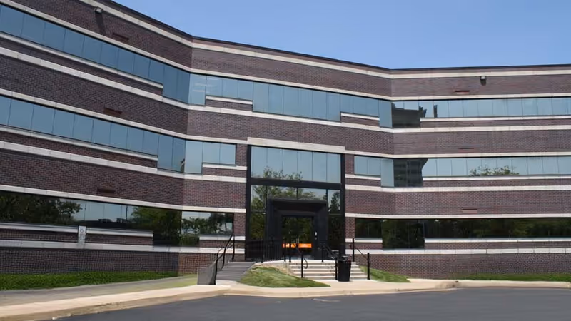 Modern brick building with glass windows and a central entrance, fronted by a paved driveway.
