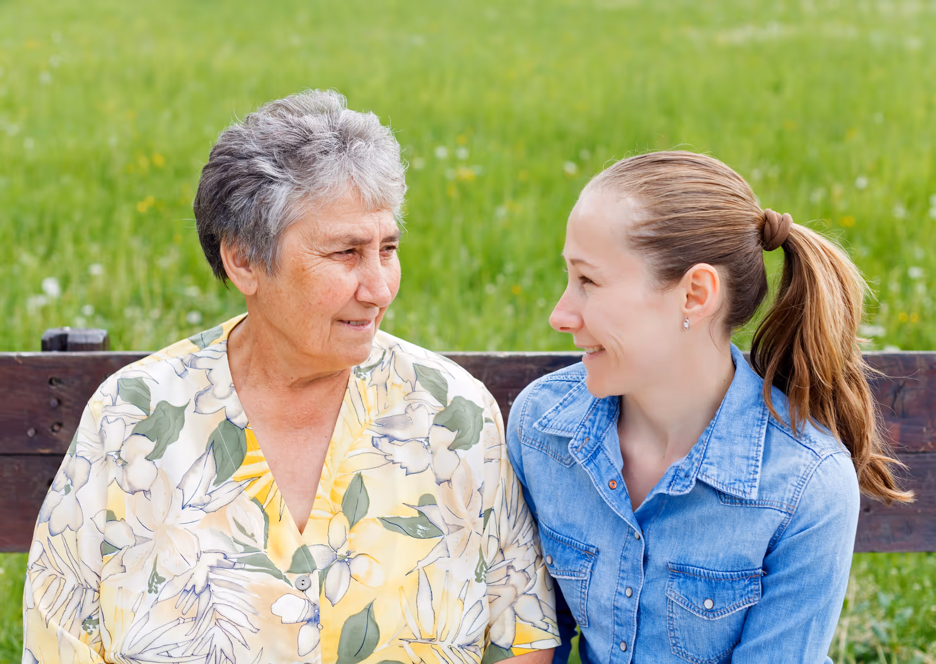 Elderly woman and caregiver chat on a bench in a green outdoor area.