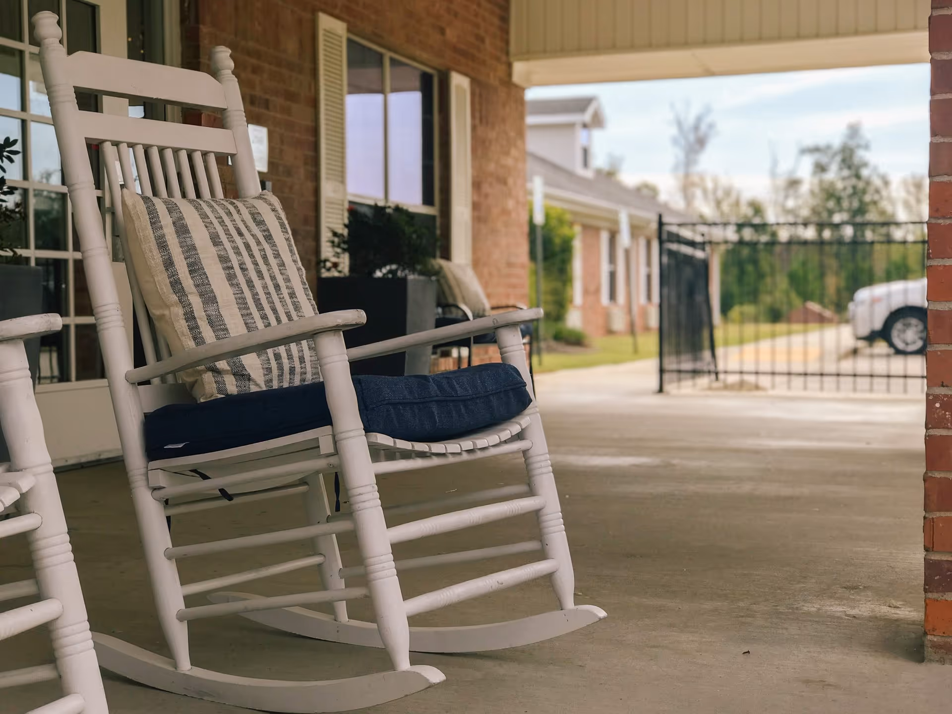 White wooden rocking chairs with cushions on a covered porch at the front of a brick senior living facility.
