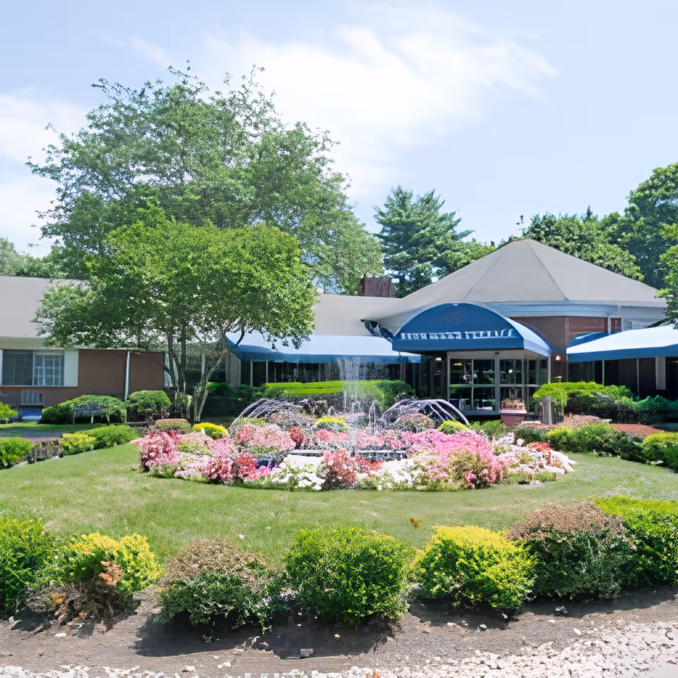 Front entrance of a senior living building with a central flowerbed and fountain, awnings, and landscaped trees and shrubs.