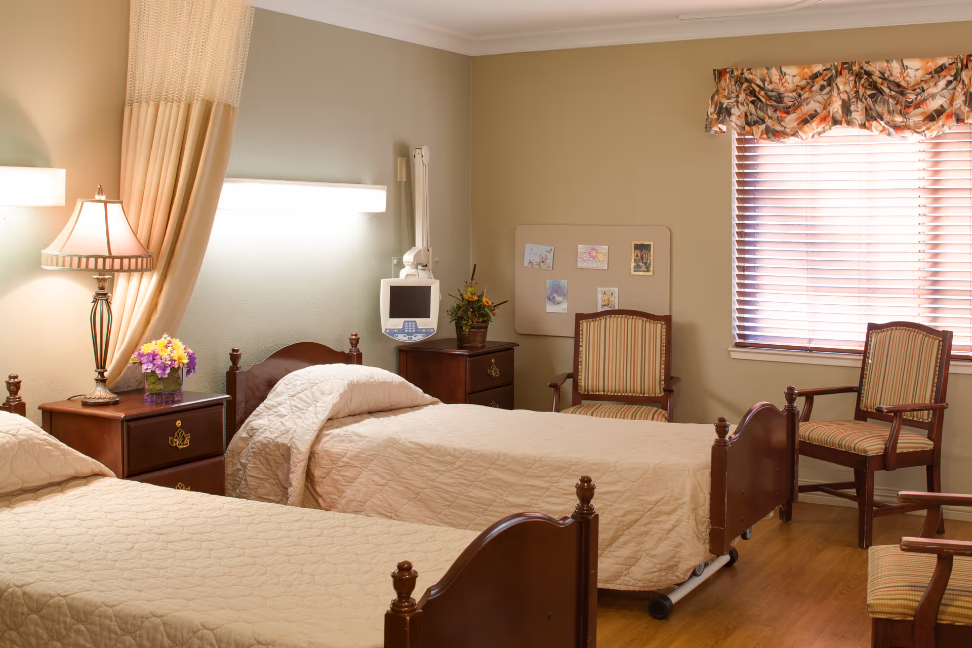 A tidy resident bedroom with two single beds, wooden nightstands, a lamp, and chairs by a window.