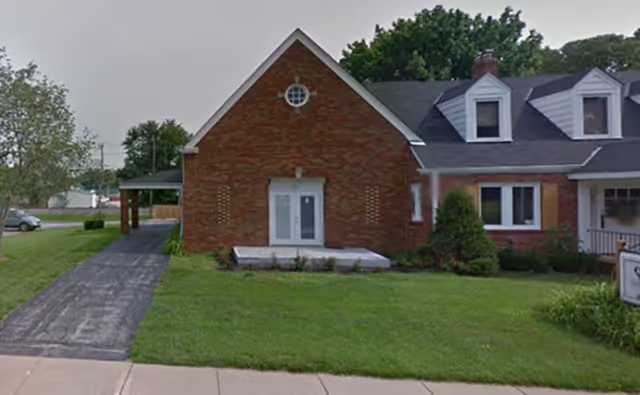 Front exterior view of a brick building with a triangular roof section, white double doors, and several windows. There is a driveway on the left side and a well-maintained lawn in front.