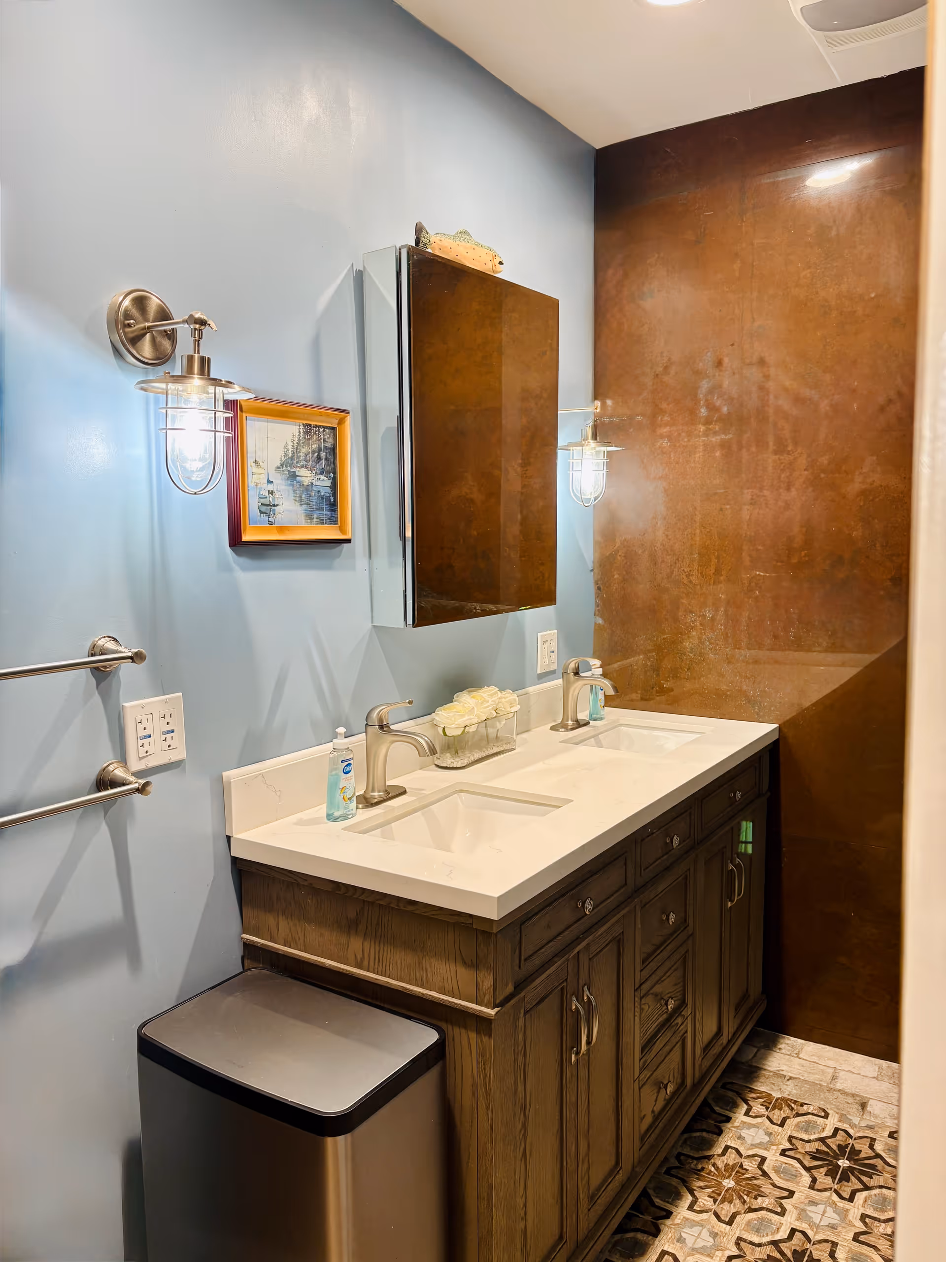 Blue-walled bathroom with double sink vanity, wall lights, and brown tiled shower alcove.