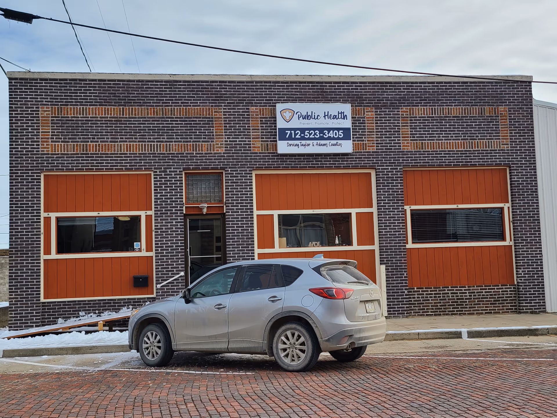Public health building with brick exterior, orange doors, and a silver SUV parked outside.