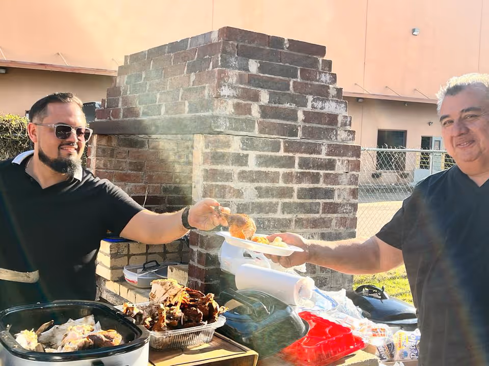 Two men barbecue outdoors by a brick grill, serving food on plates.