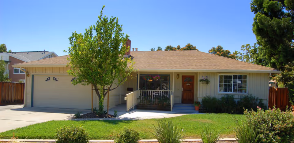 Front exterior of a single-story suburban house with an attached two-car garage, a small porch, a tree, and a manicured lawn.